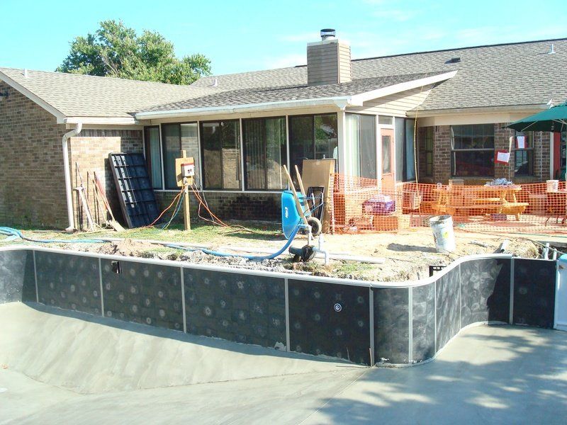 Pool under construction near a house; black walls, equipment, and patio furniture.