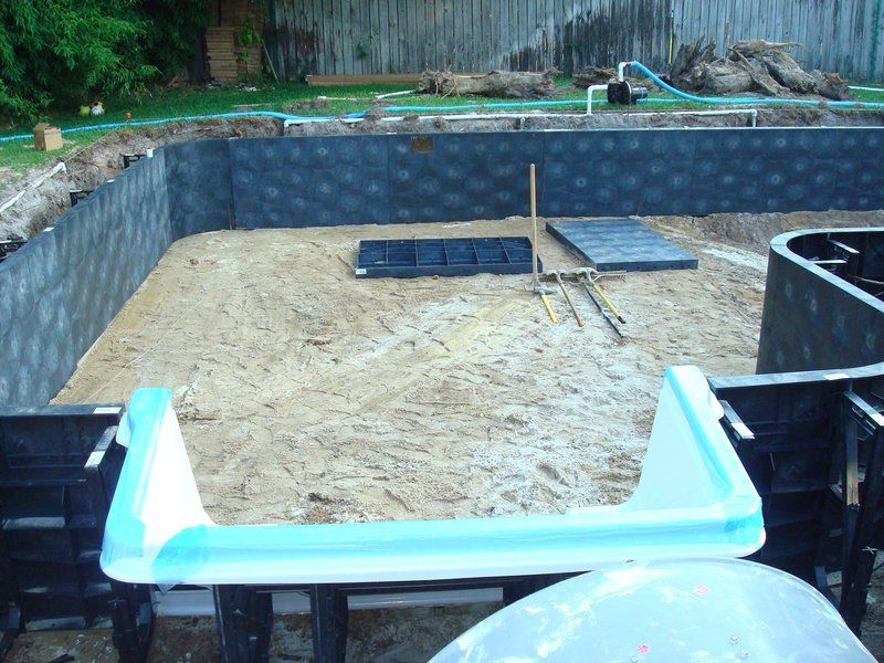 Pool construction: Excavated hole with sand base, black walls, and equipment. Blue coping in foreground.