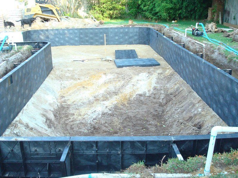 Pool construction site. Excavated hole with dark walls, dirt floor, piping, and machinery.