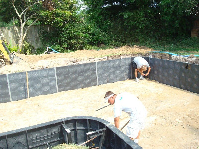 Pool construction: Two workers inside a pool foundation, installing black wall panels, sandy ground.