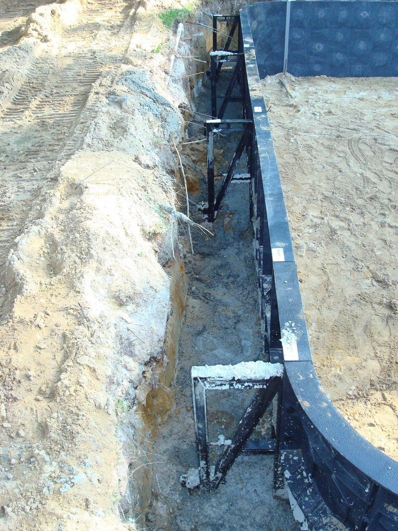 Construction of a curved pool wall, supported by metal bracing, in an excavated trench. Brown soil surrounds it.