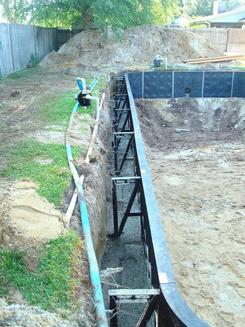 Pool construction site: Excavated earth, retaining walls with supports, blue hose, pump.