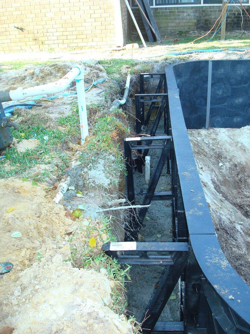 Construction of a curved pool frame next to a house; black metal structure with plumbing and dirt.