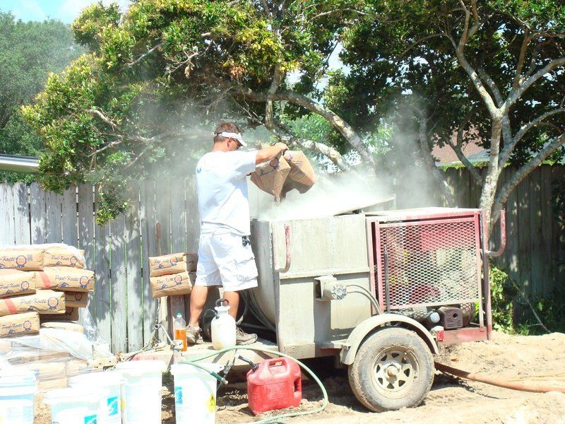 Man adding cement to a cement mixer outdoors, creating a cloud of dust near a fence.