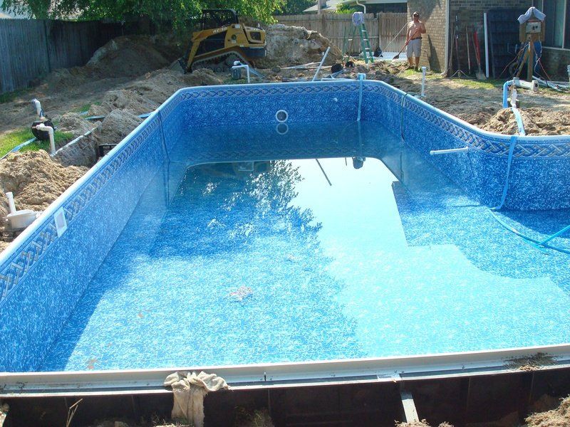 Partially constructed swimming pool with blue mosaic-patterned liner and water, surrounded by dirt and construction equipment.