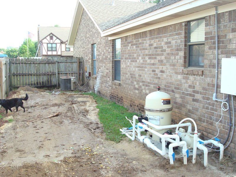Backyard with pool equipment by a brick house; a black dog walks on the dirt.