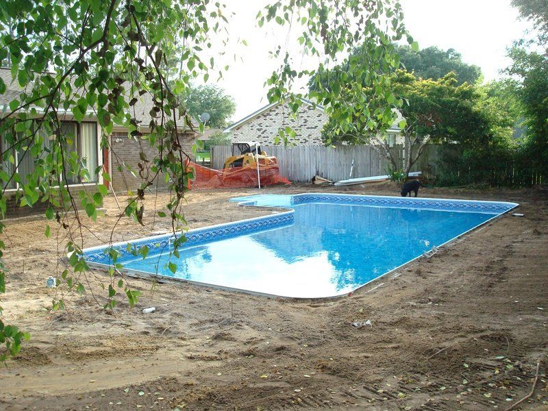 Newly constructed rectangular swimming pool with blue tile interior. Dirt surrounds the pool; a house and trees are in the background.