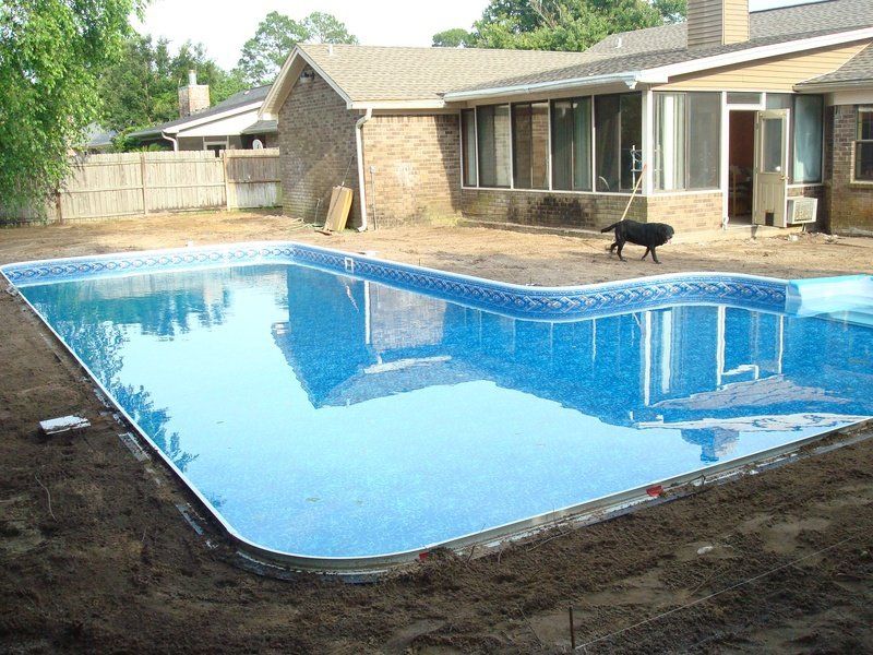 A rectangular pool filled with blue water next to a brown brick house. A dog is near the house.