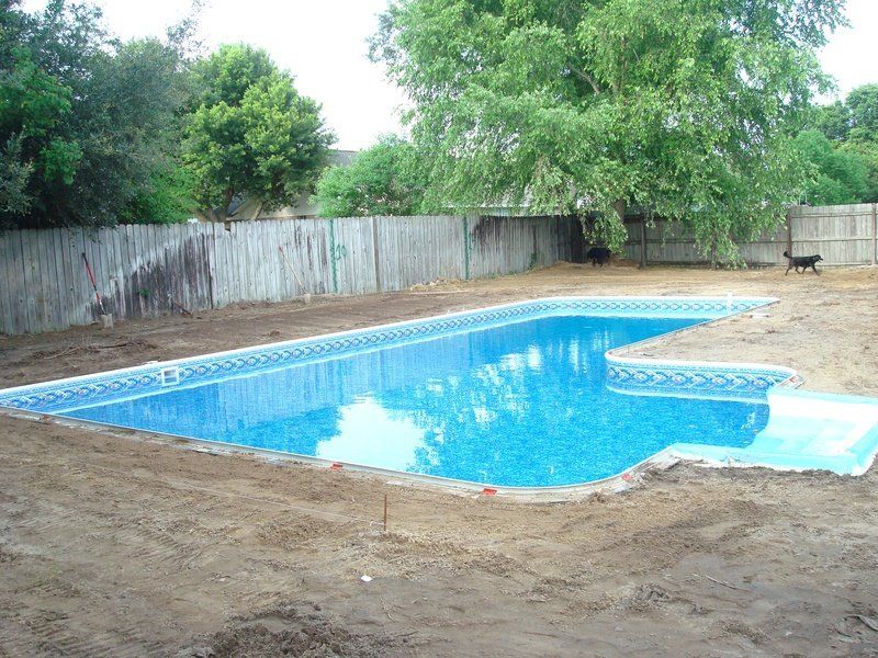 Rectangular swimming pool with blue water, surrounded by dirt, near a wooden fence and trees.