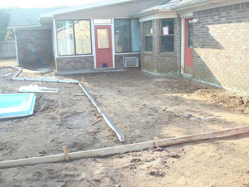 Backyard construction site; concrete forms in dirt, beside a brick building with a red door and a screened-in porch.