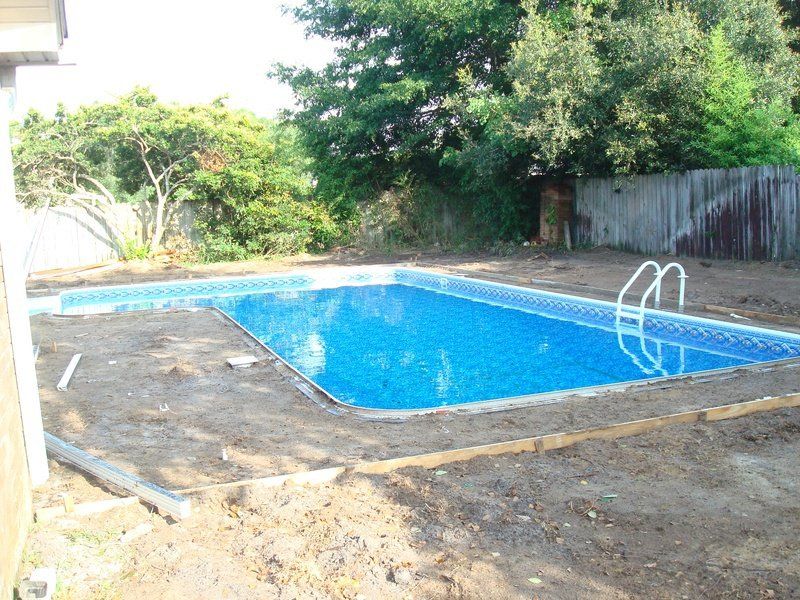 Rectangular in-ground pool filled with blue water, surrounded by dirt and a wooden border.