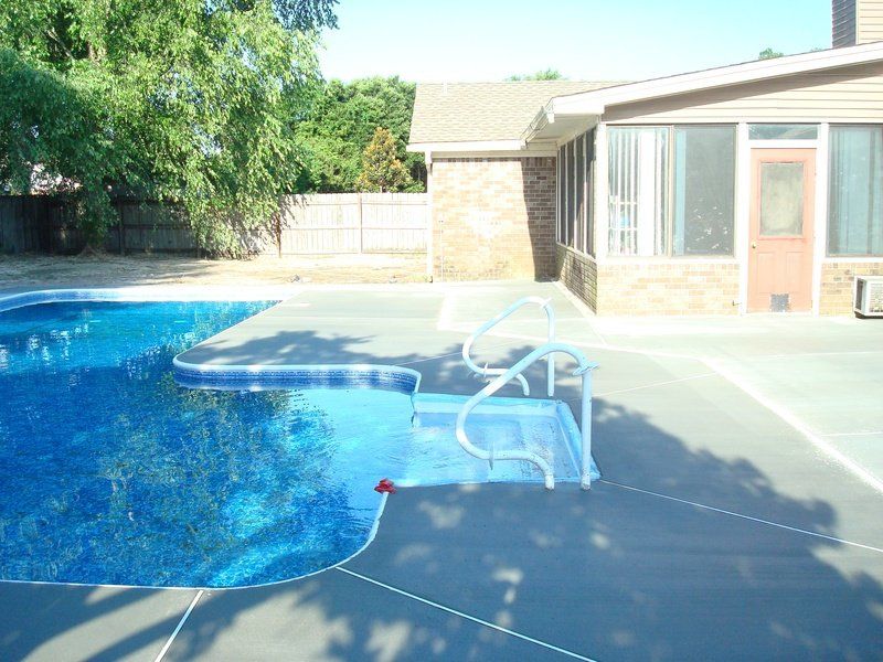 Pool with blue water and gray concrete surrounding it. House visible in the background.