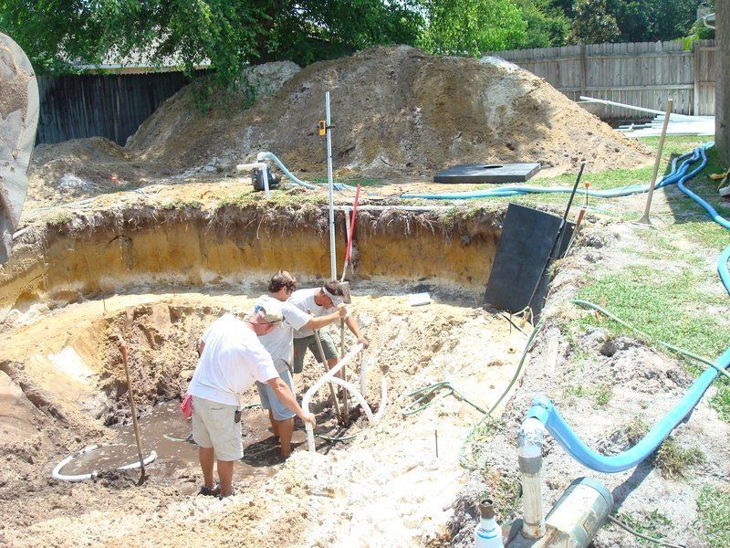 Men working to excavate a pool. Dirt pile, water hoses, and surveying equipment visible.