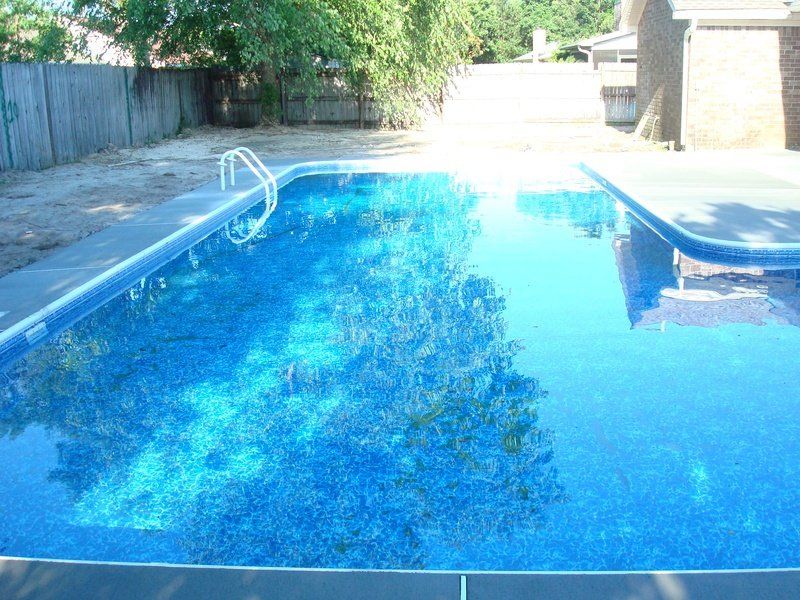 A rectangular swimming pool filled with water, trees reflected on surface, steps with a rail in the pool.