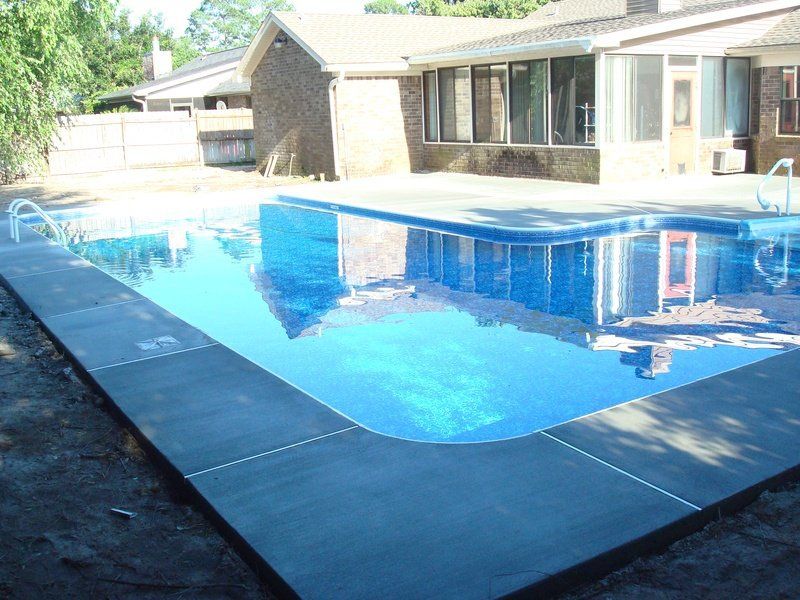 Rectangular swimming pool with blue water and gray concrete edging, next to a brick house.