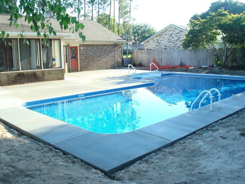 Rectangular swimming pool with blue water and gray concrete edging, next to a brick building.