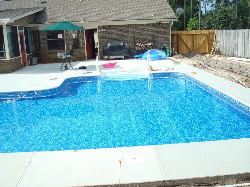 Swimming pool with blue water surrounded by concrete patio; backyard setting.