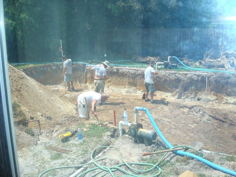 Construction workers excavating a backyard pool. Dirt piles, blue water hoses, and a fence visible.