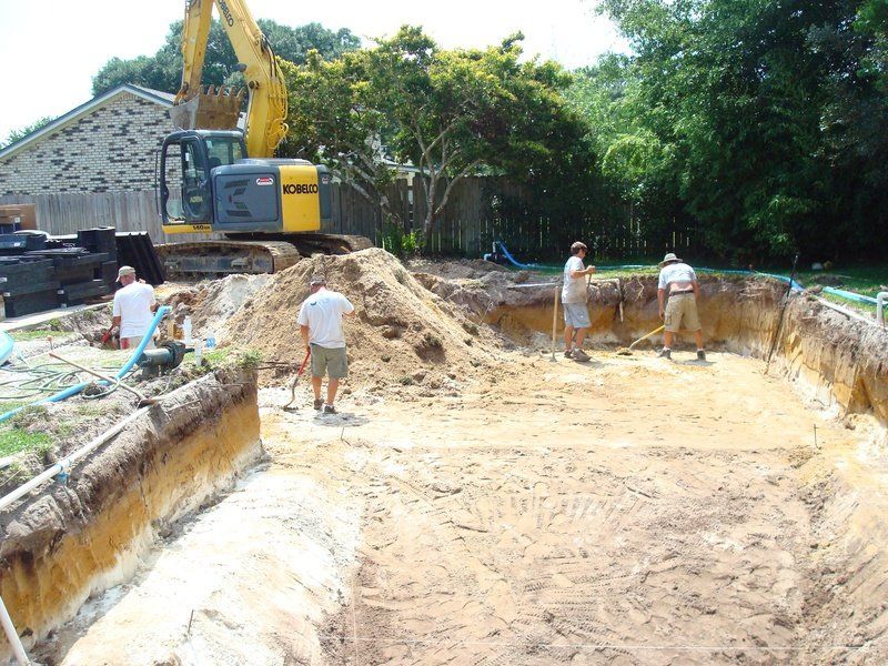 Pool excavation in progress: excavator, workers leveling soil, dirt pile, and supplies in yard.