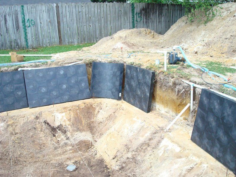 Pool construction: black wall panels installed in a sand pit, with pipes and dirt pile visible.