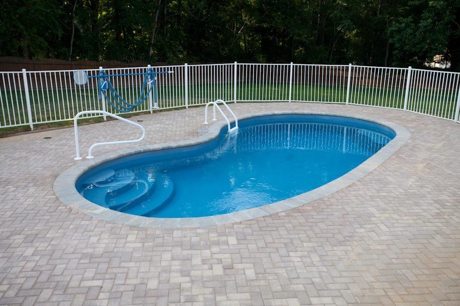 Blue kidney-shaped swimming pool surrounded by brick pavers and a white fence.