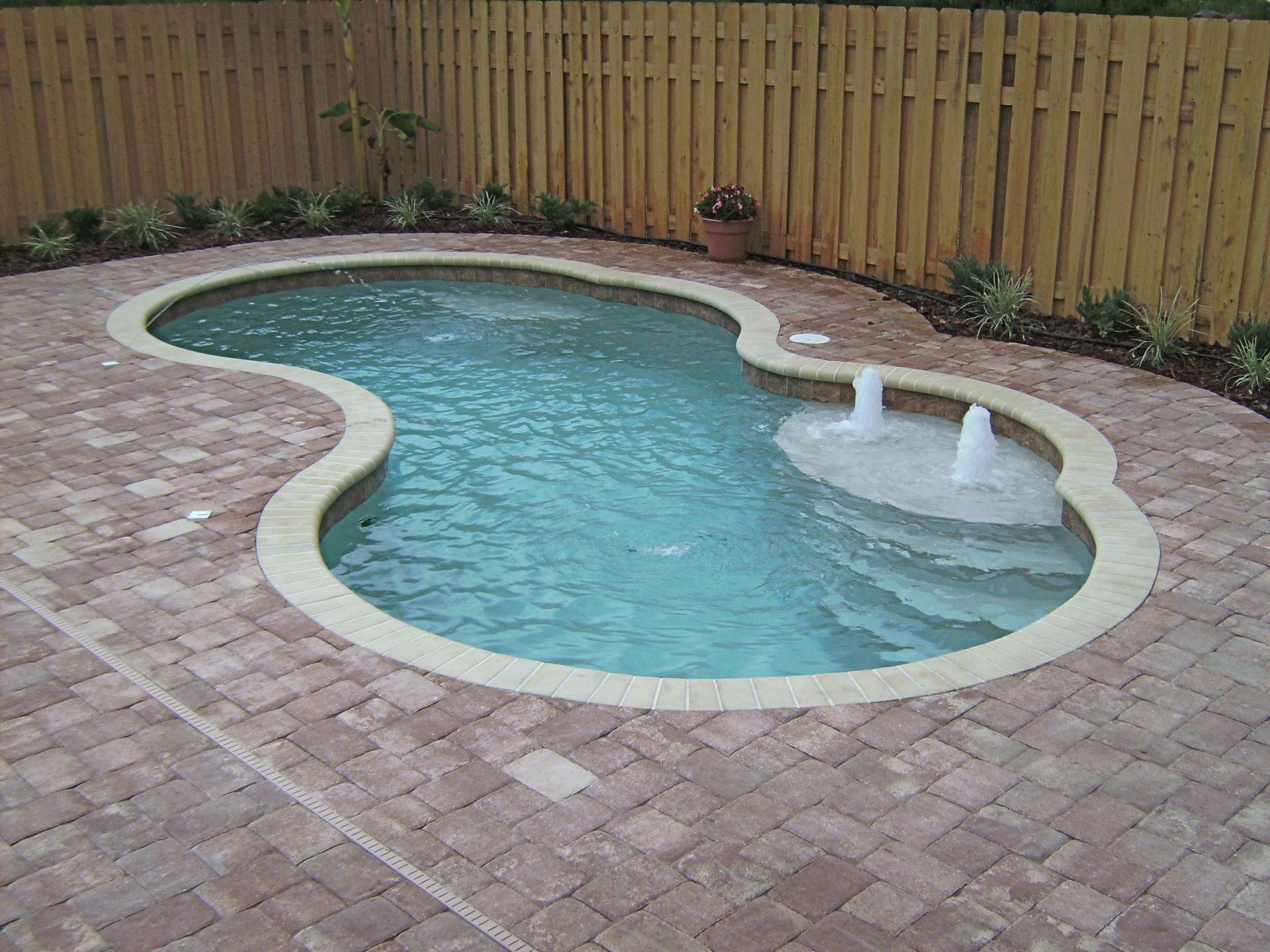 Pool with brick patio and fountain. Surrounded by wooden fence and greenery.