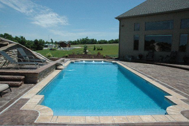 Rectangular in-ground pool with blue water and stone patio, adjacent to a two-story house, sunny day.