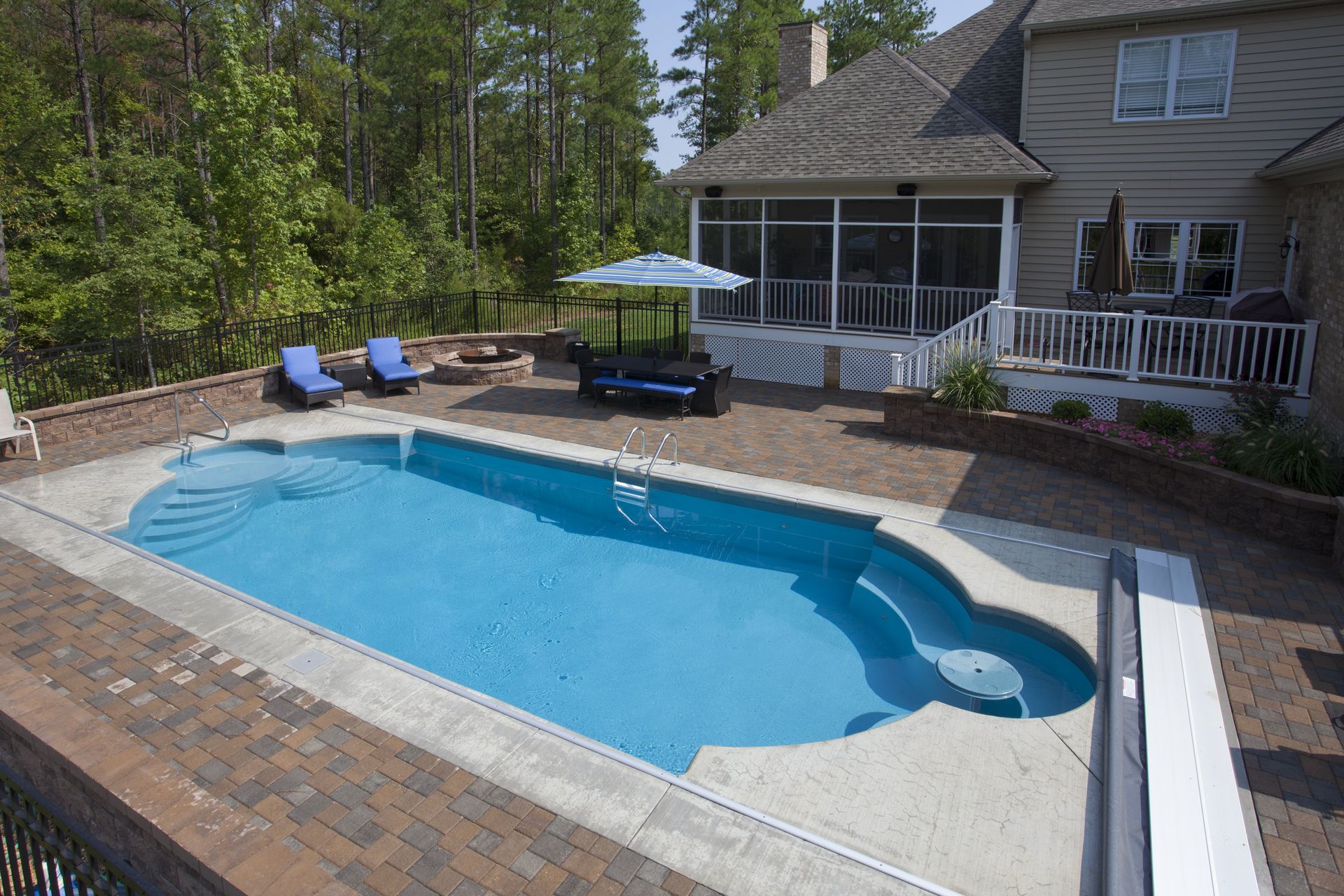 Backyard pool with surrounding brick patio, two lounge chairs, and a house in the background.