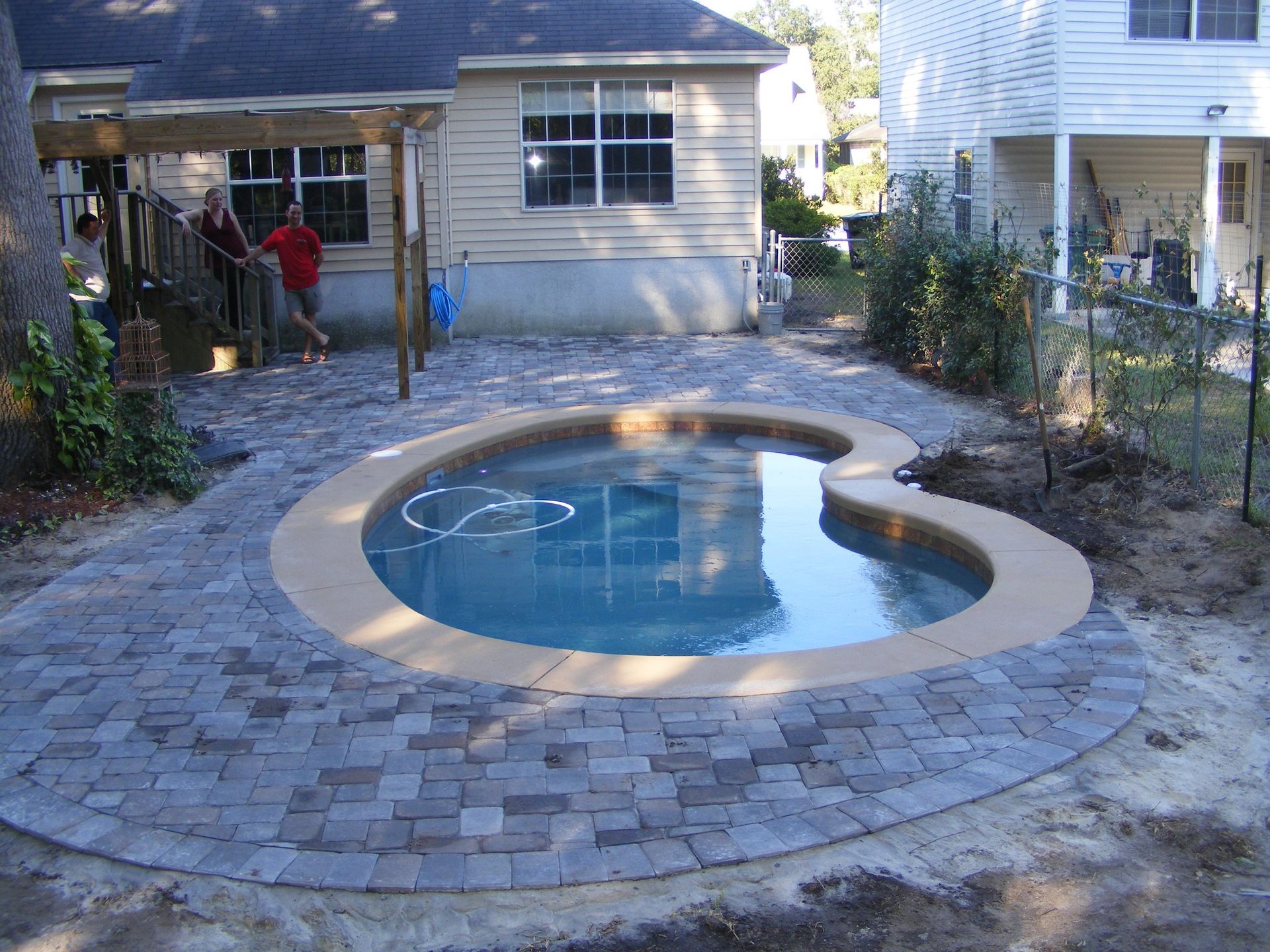 Pool surrounded by gray pavers. Two people on a deck behind the pool. Houses and a fence are in the background.