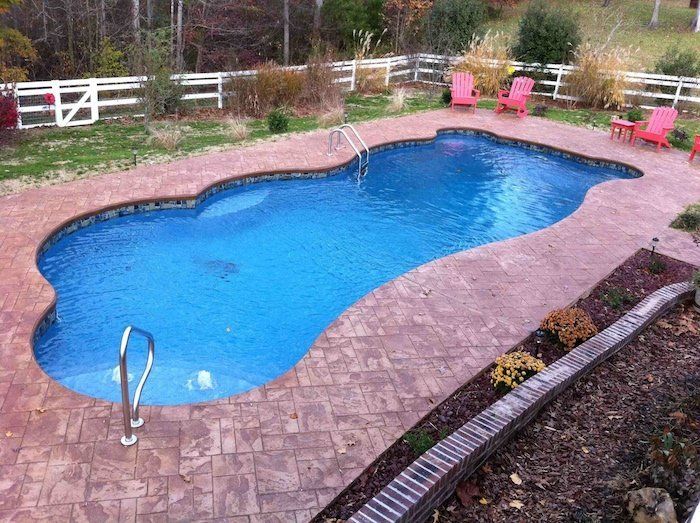 A kidney-shaped swimming pool with blue water, surrounded by red brick, with red chairs and a white fence in the background.