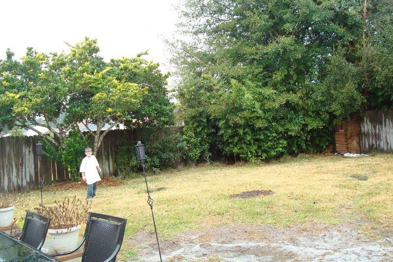 Person in a white shirt in a grassy backyard, surrounded by trees and a wooden fence.