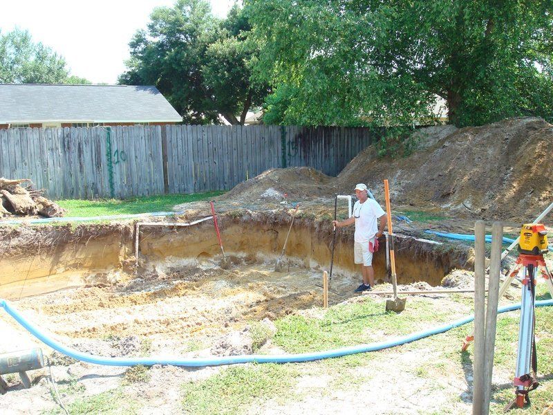 Man standing in excavated area with dirt piles, blue pipes, and fencing in background.