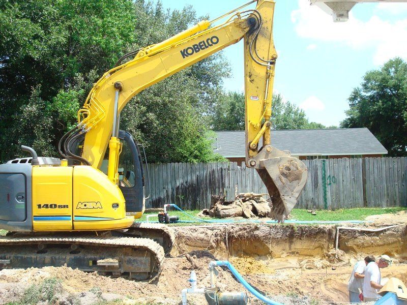 Yellow Kobelco excavator digging in a dirt pit near a wooden fence and house.