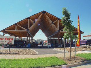 Farmers market under a wooden, geometric roof. Vendors and shoppers present. Bright, sunny day.
