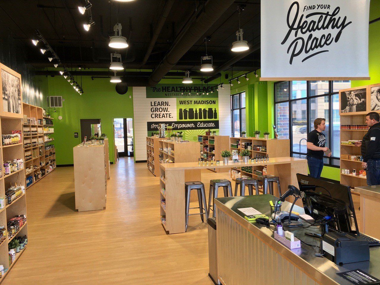 Interior of a bright, green-accented health food store. Display shelves, customers, and a sign that says 