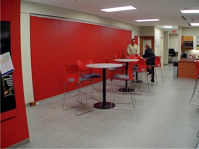 Red wall with cafe tables and chairs. Two people sit at a table. Fluorescent lights.