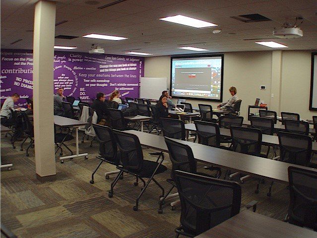 Classroom with people using laptops, projector screen displaying a graph, purple wall, and rows of chairs.