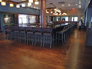 Bar with long counter and stools, wooden floor, lighting fixtures, and a central pillar.