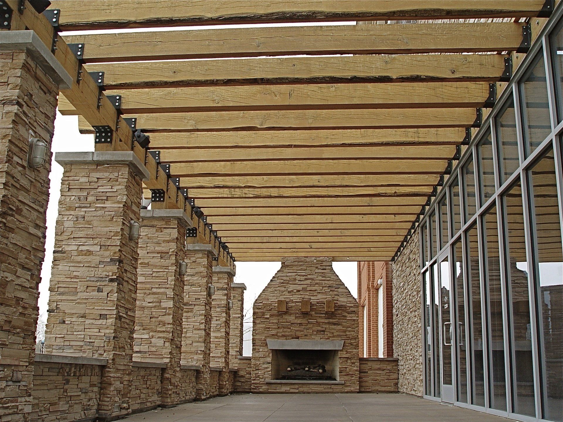 Stone columns and a pergola cover an outdoor patio with a fireplace and glass wall.