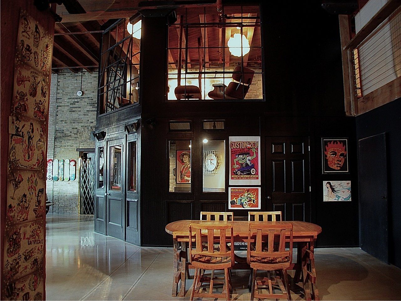 Interior of an art gallery with dark walls, wooden table, chairs, and displayed posters.