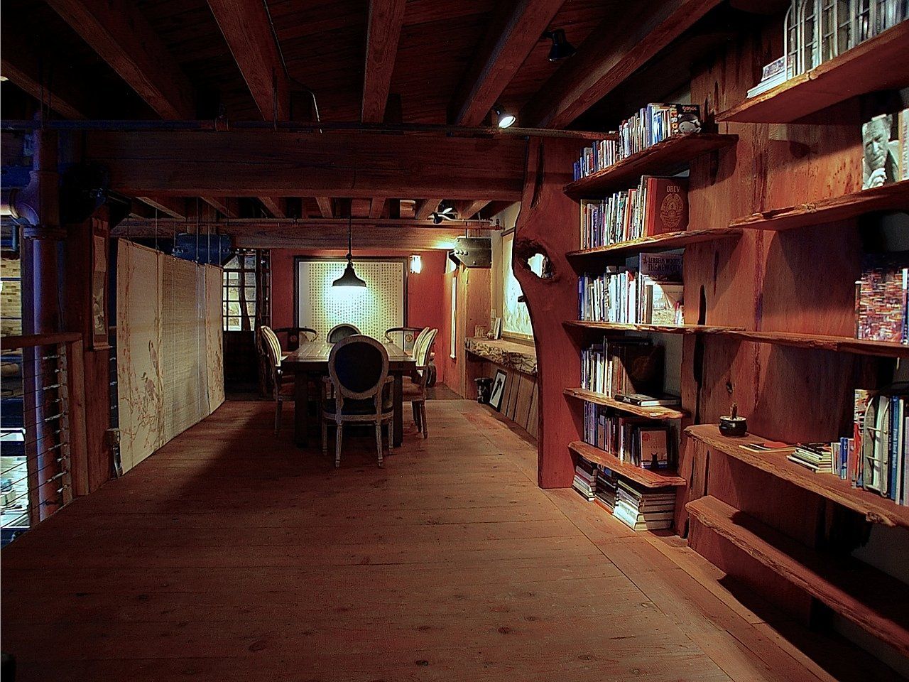 Wooden library with bookshelves lining one wall, dining table with chairs, and overhead beams.