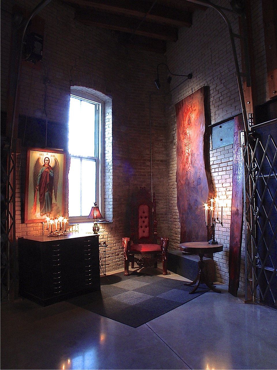 A dimly lit room with a window, chair, artwork, and various furniture against brick walls.