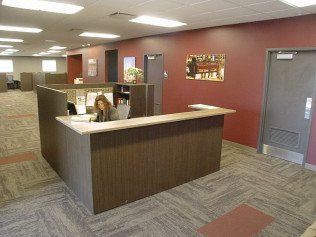 Office reception area: Person at desk. Red and neutral-toned walls, cubicles in background.