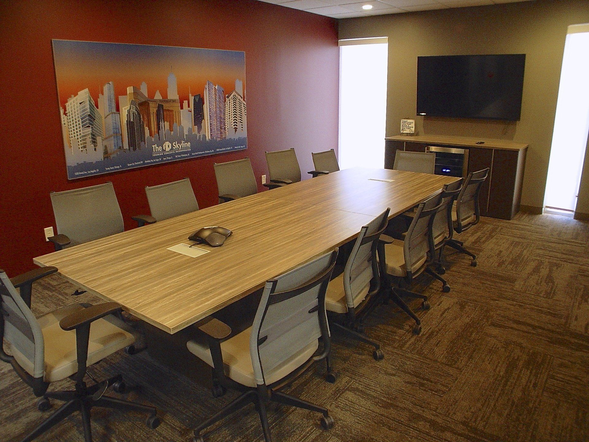 Conference room with long table, chairs, and cityscape artwork on red wall. A TV is mounted on a brown cabinet.