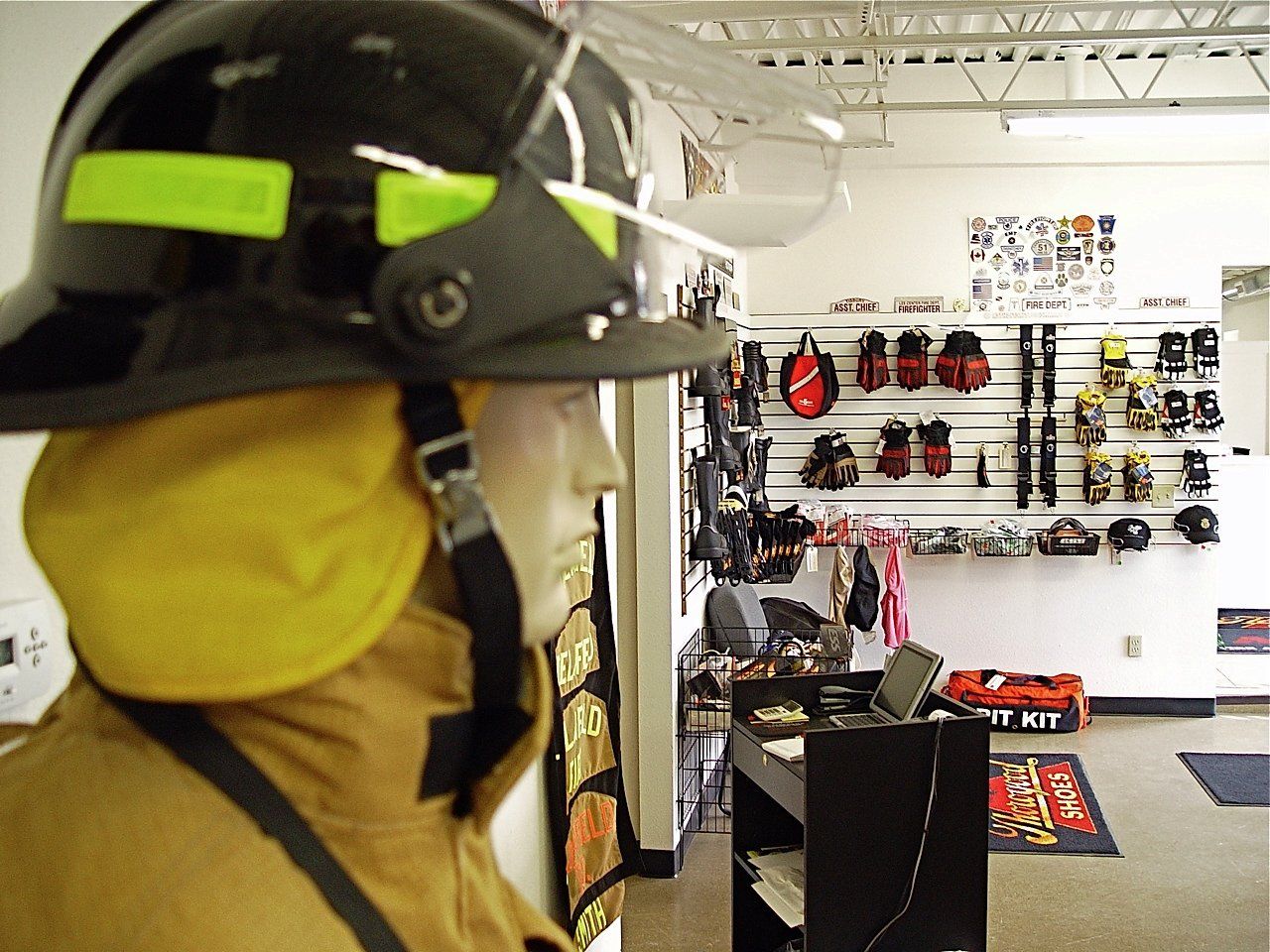 Mannequin in firefighter gear, helmet and coat, in a fire equipment store, with gear displayed in the background.
