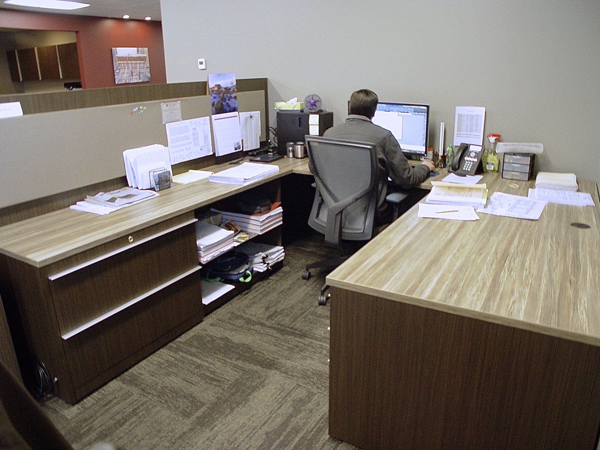 Man working at a desk in an office. Wooden desk with computer, papers, and files. Brown carpet.