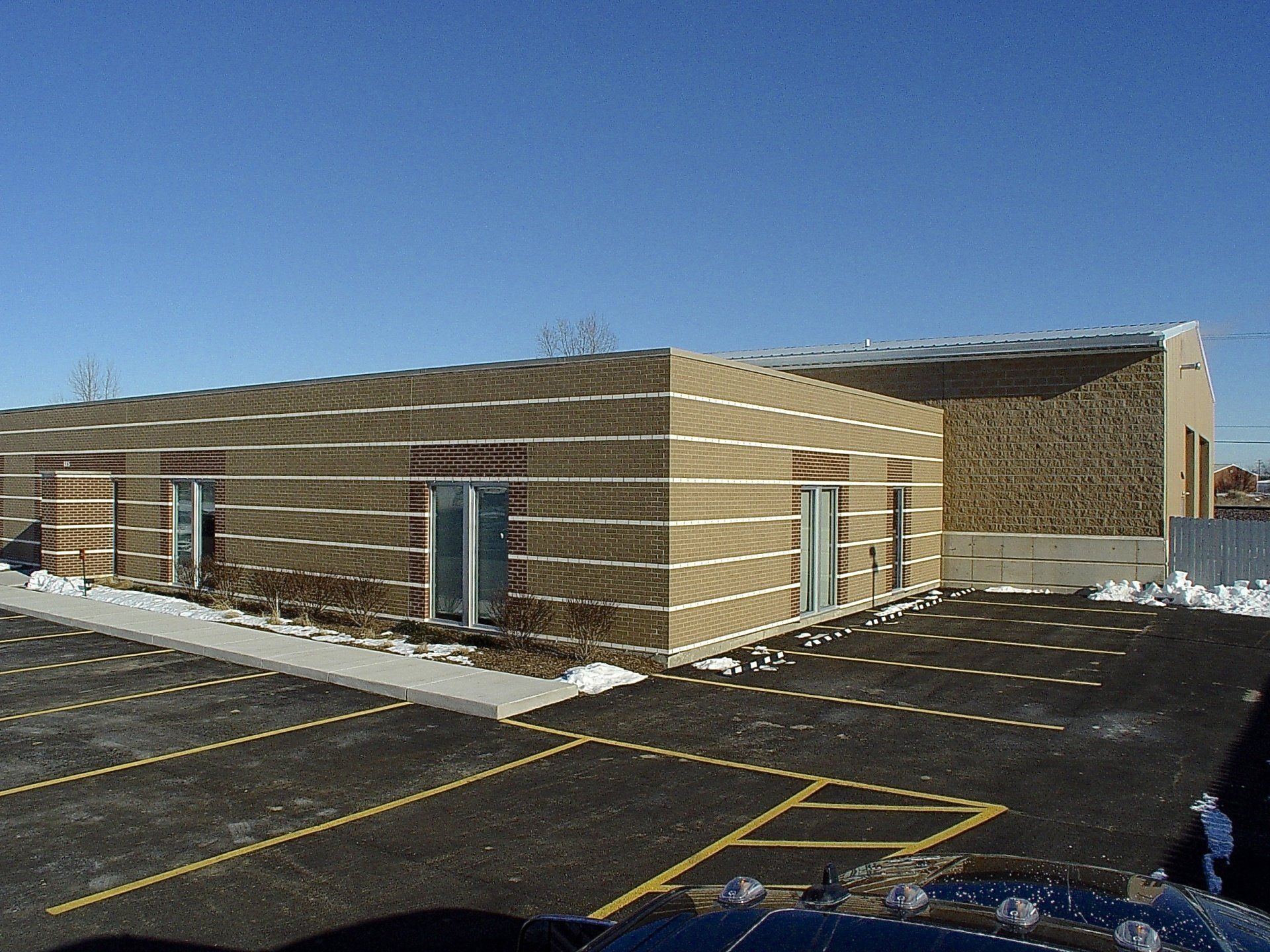 Exterior view of a one-story building with tan siding, windows, and a parking lot on a sunny day.