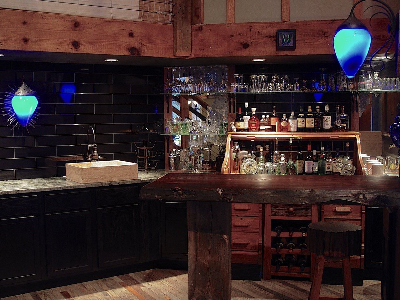 Dark bar with wood counter and cabinets, lit by blue glowing lights and shelves of liquor bottles.
