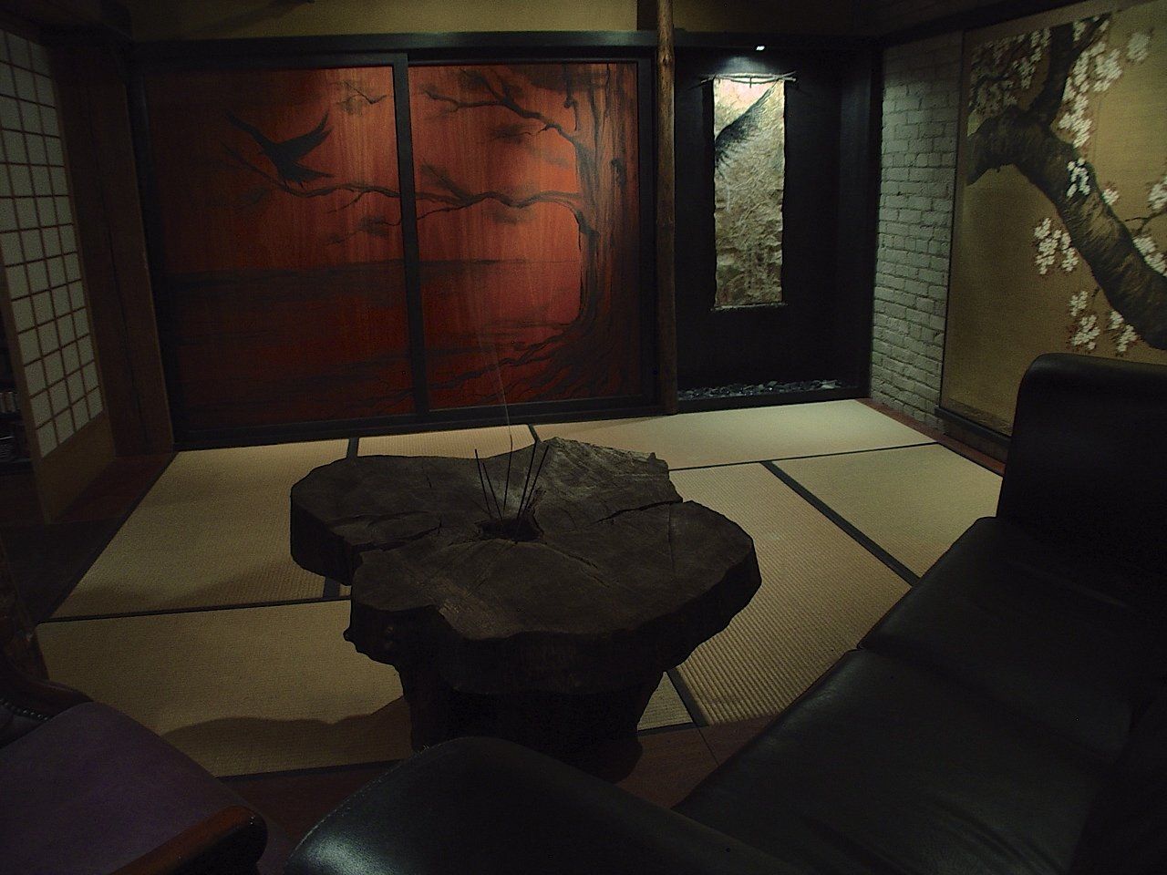 Cozy Japanese-style room with a tree-trunk table, tatami mats, a red-paneled screen, and a black leather chair.
