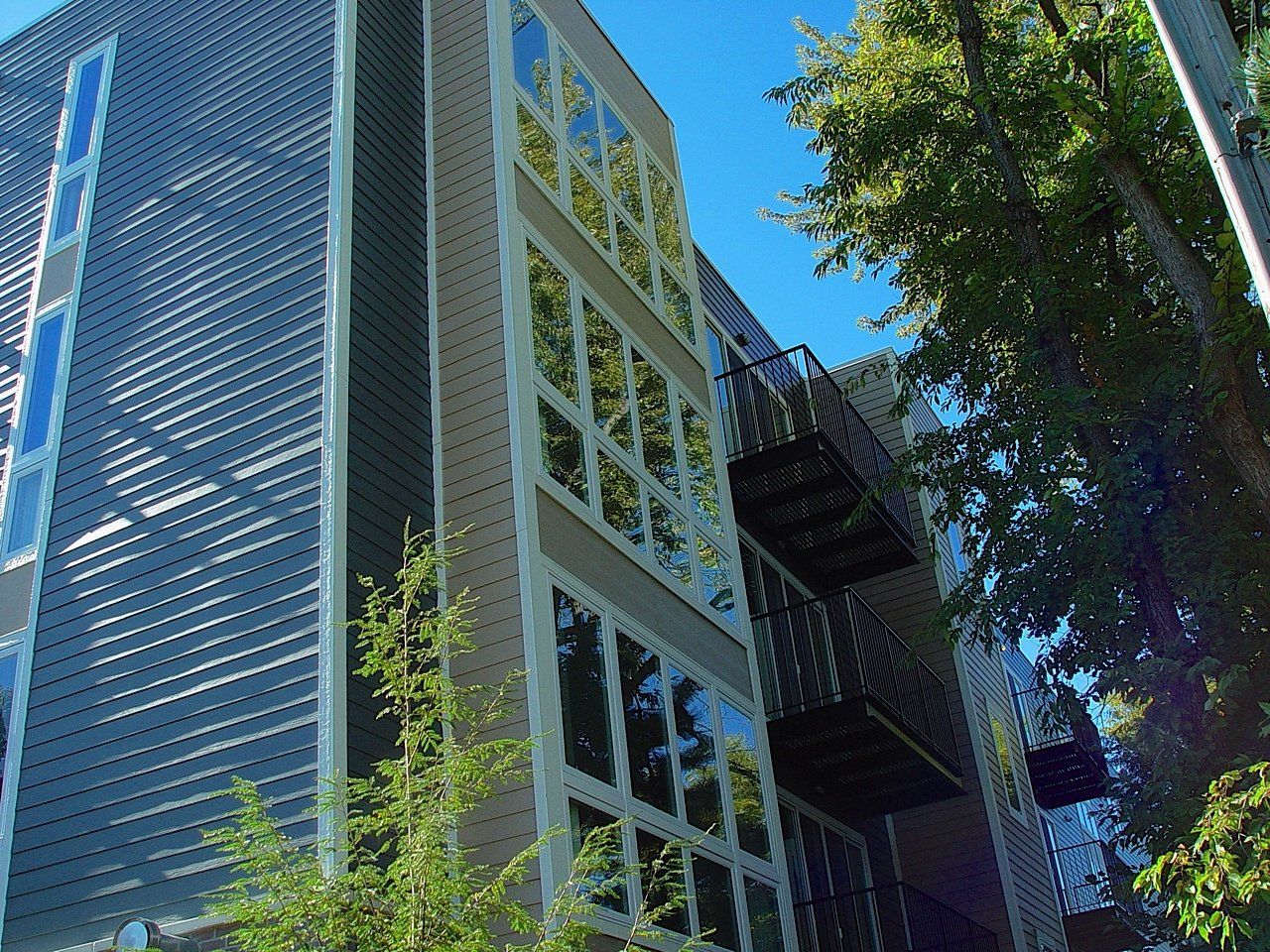 Modern multi-story building with large windows and metal facade, with balconies, trees, and blue sky.